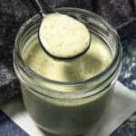 A spoon holds creamy, greenish Jalapeno Ranch Dressing above a glass jar filled with the same sauce, sitting on a white napkin with a dark textured background.