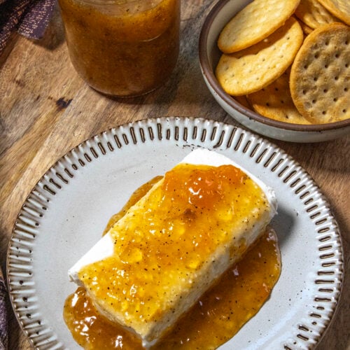 A plate with cream cheese topped with golden, zesty Jezebel Sauce, next to a bowl of round crackers and a jar of jezebel sauce on a wooden surface.