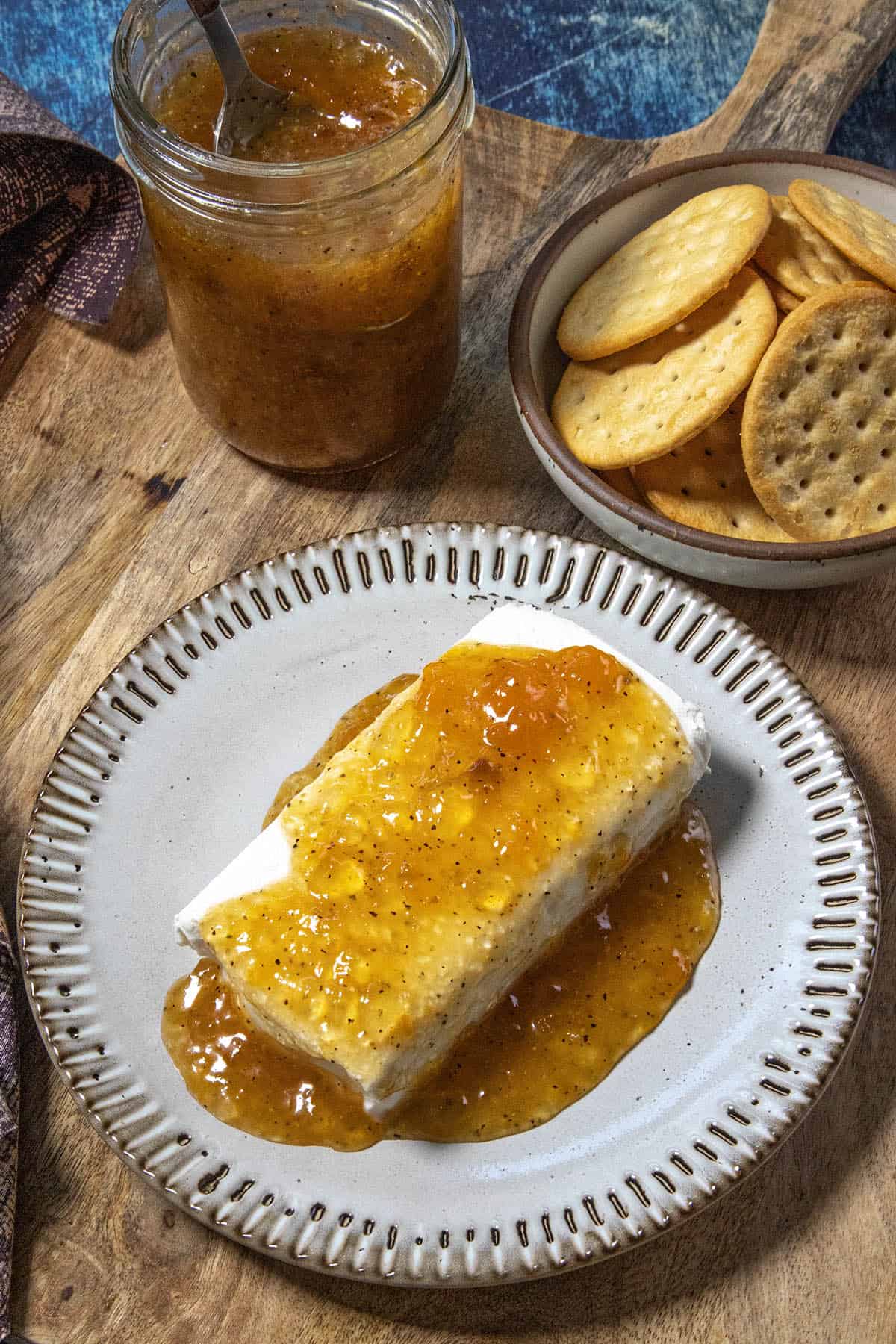 A plate with cream cheese topped with golden, zesty Jezebel Sauce, next to a bowl of round crackers and a jar of jezebel sauce on a wooden surface.