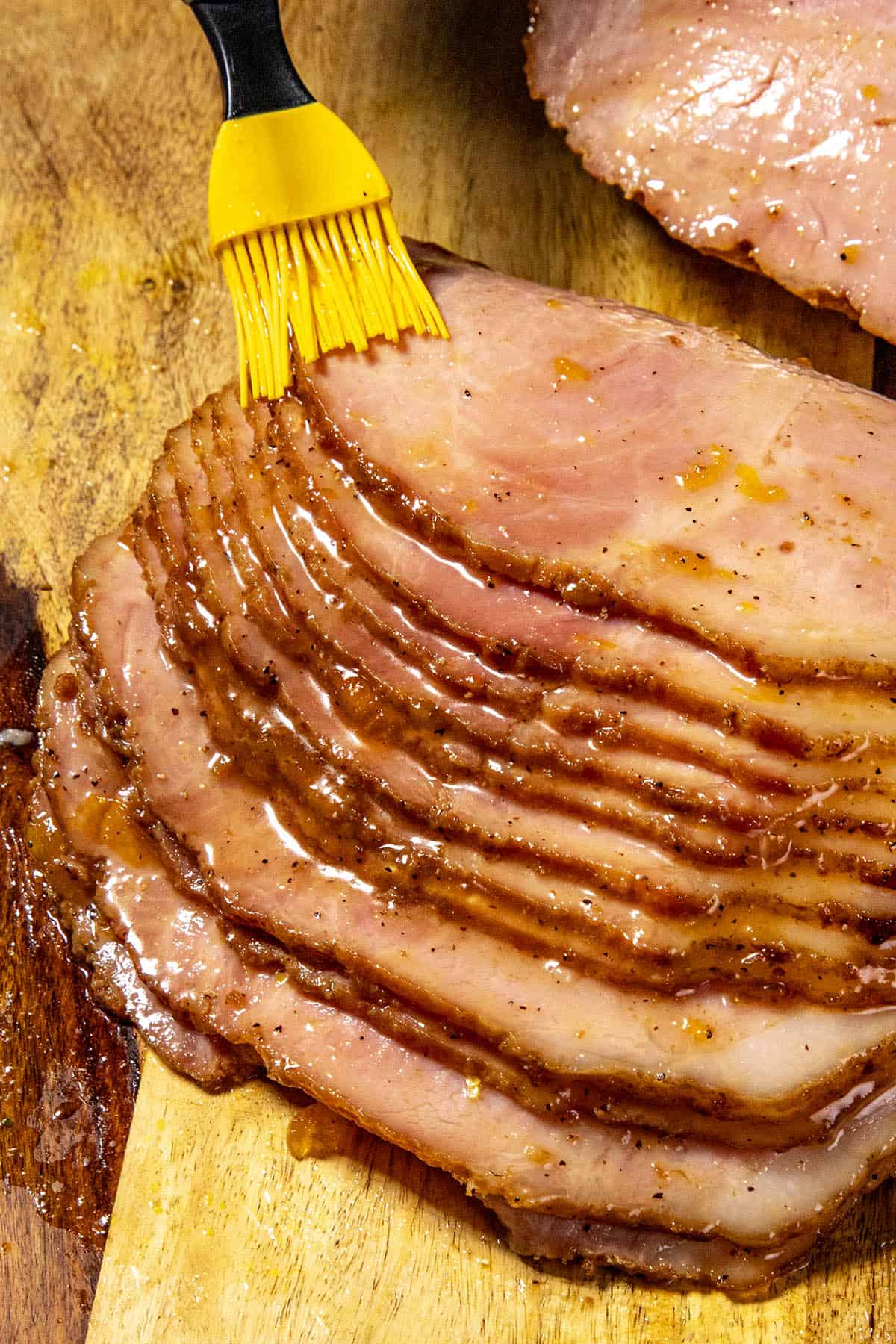 A yellow brush spreads a generous layer of Jezebel Sauce glaze over sliced ham on a wooden cutting board, highlighting the juicy, caramelized surface of the meat.