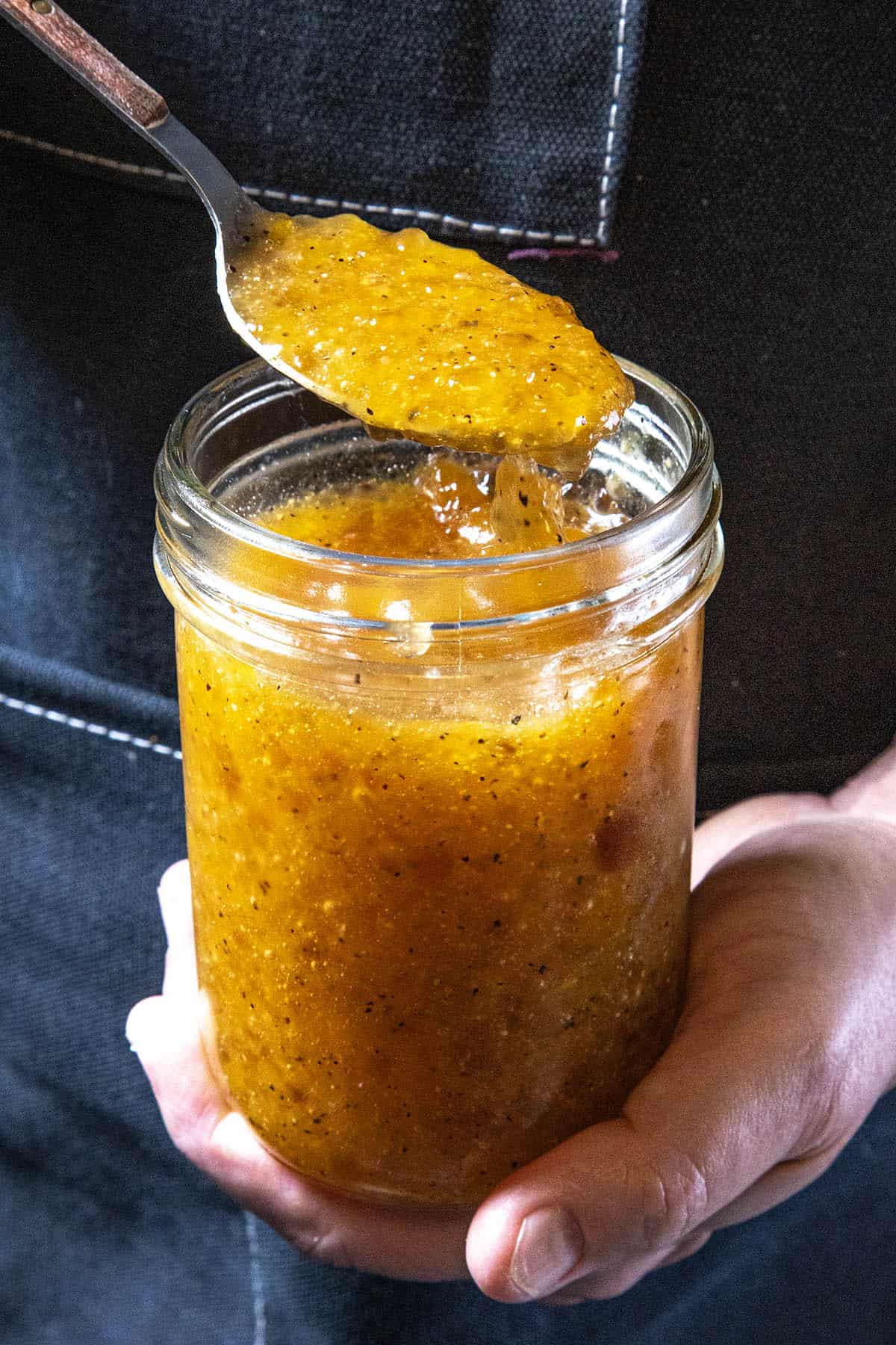 Mike from Chili Pepper Madness holds a glass jar filled with yellow homemade jam or marmalade—perhaps even a tangy Jezebel Sauce—and lifts some out with a spoon.
