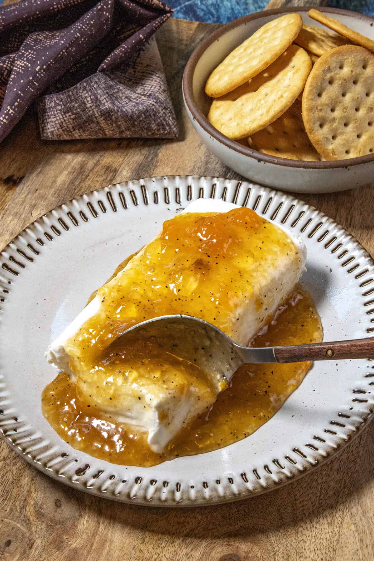 A block of cream cheese topped with golden-orange Jezebel Sauce sits on a plate. A spoon rests on it, spreading the sweet and tangy sauce. In the background, a bowl of round crackers is visible on a wooden surface.