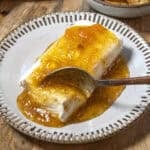 A block of cream cheese is topped with golden, chunky Jezebel Sauce on a plate, with a spoon scooping some of the sweet and tangy sauce along with the creamy cheese.