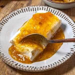 A block of cream cheese is topped with golden, chunky Jezebel Sauce on a plate, with a spoon scooping some of the sweet and tangy sauce along with the creamy cheese.