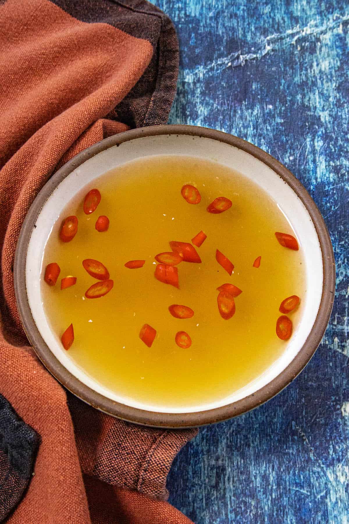 A bowl of golden Nuoc Cham (Vietnamese Dipping Sauce) with sliced red chili peppers sits on a blue surface next to a rust-colored cloth.