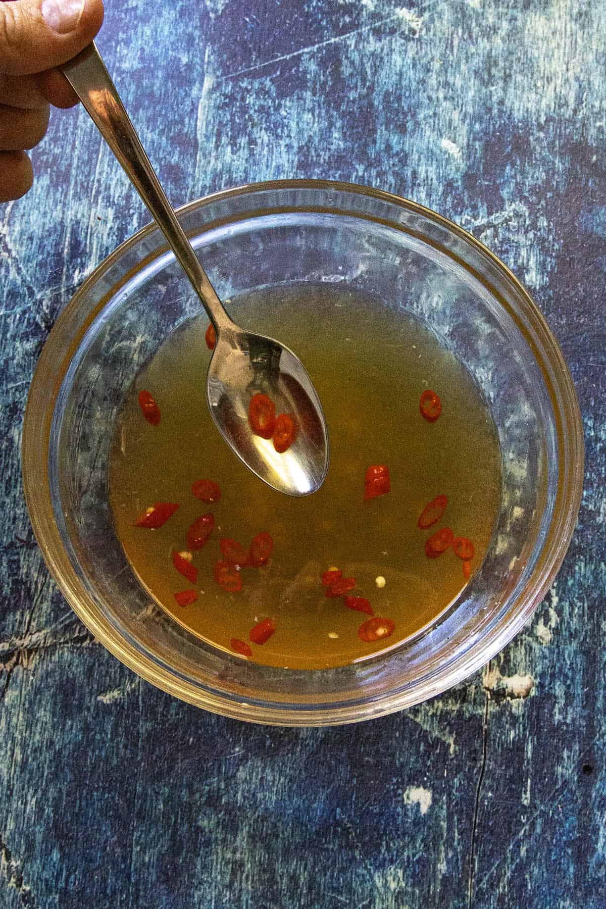 A hand holds a spoon above a glass bowl filled with light brown Nuoc Cham and sliced red chili peppers, set on a blue textured surface.