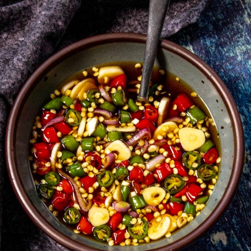A bowl of chopped red and green chilies, garlic slices, and onions sits in a dark liquid—classic Prik Nam Pla (Thai Chili Fish Sauce)—with a spoon inside, placed on a textured dark cloth and blue surface.