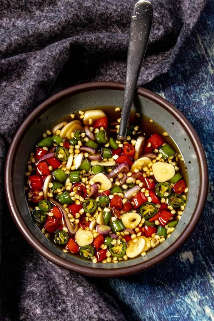 A bowl of chopped red and green chilies, garlic slices, and onions sits in a dark liquid—classic Prik Nam Pla (Thai Chili Fish Sauce)—with a spoon inside, placed on a textured dark cloth and blue surface.