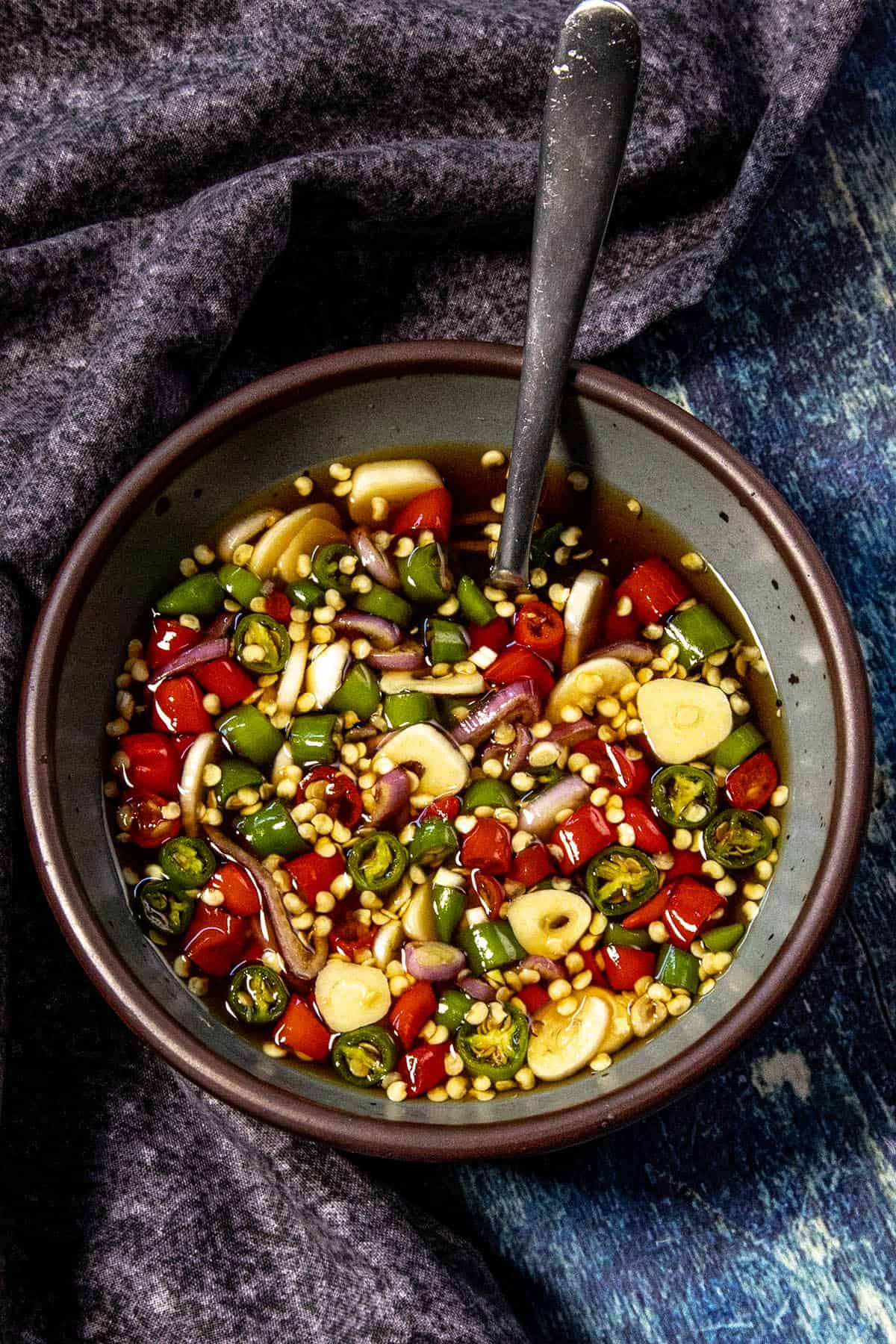 A bowl of chopped red and green chilies, garlic slices, and onions sits in a dark liquid—classic Prik Nam Pla (Thai Chili Fish Sauce)—with a spoon inside, placed on a textured dark cloth and blue surface.