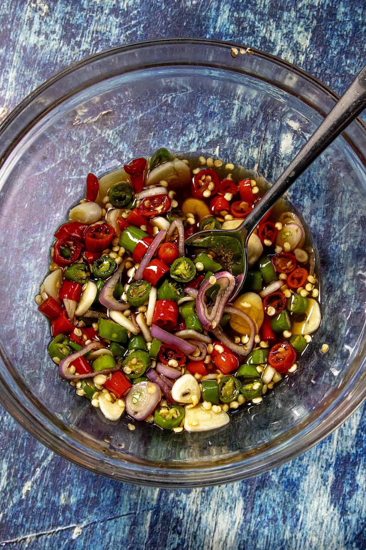 A glass bowl filled with sliced red and green chili peppers, shallot, and garlic in a clear liquid, known as Prik Nam Pla, with a spoon inside, sits on a textured blue surface.