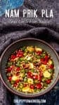 Bowl of Prik Nam Pla Thai chili fish sauce with sliced red and green chilies, garlic, and onions, sitting on a dark cloth background.