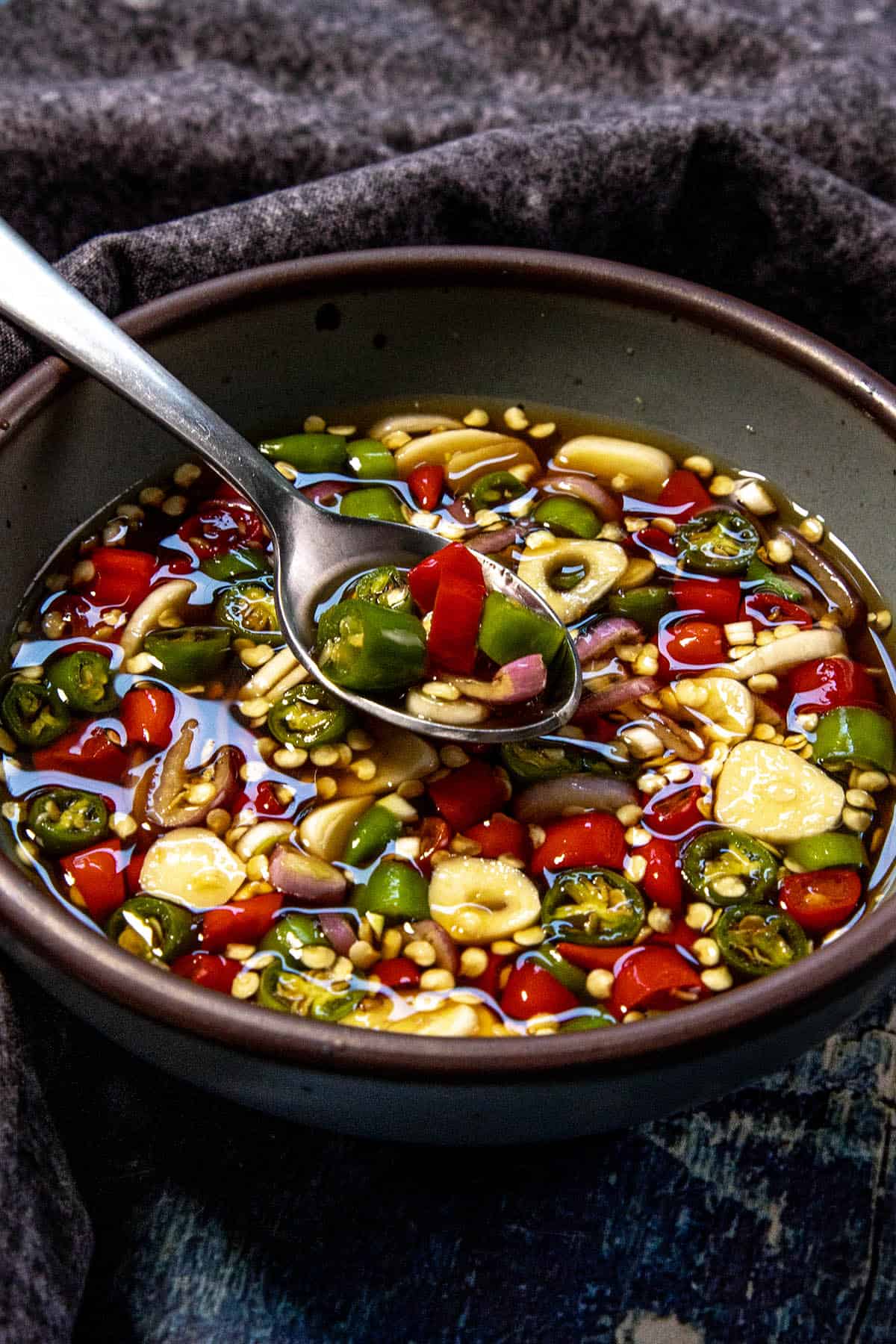 A bowl of fish sauce with sliced green and red chilies, garlic, and shallot—a classic Thai Chili Fish Sauce Condiment (Authentic Prik Nam Pla)—with a spoon lifting some of the vibrant mixture. The bowl is placed on a dark textured cloth.