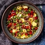 A bowl filled with sliced red and green Thai chili peppers, garlic, and seeds in fish sauce - classic Prik Nam Pla Recipe (Thai Chili Fish Sauce) - is placed on a gray plate with a dark textured cloth beside it.
