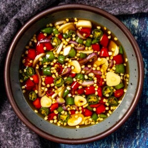 A bowl filled with sliced red and green Thai chili peppers, garlic, and seeds in fish sauce - classic Prik Nam Pla Recipe (Thai Chili Fish Sauce) - is placed on a gray plate with a dark textured cloth beside it.
