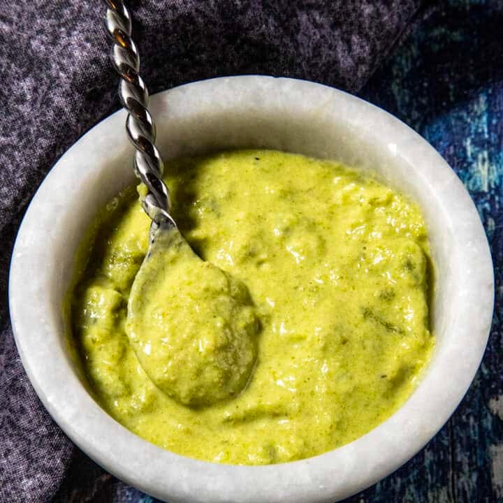 A marble bowl filled with thick green Spicy Roasted Jalapeño Sauce or chutney, with a twisted metal spoon resting inside, placed on a dark fabric napkin and a blue textured surface.
