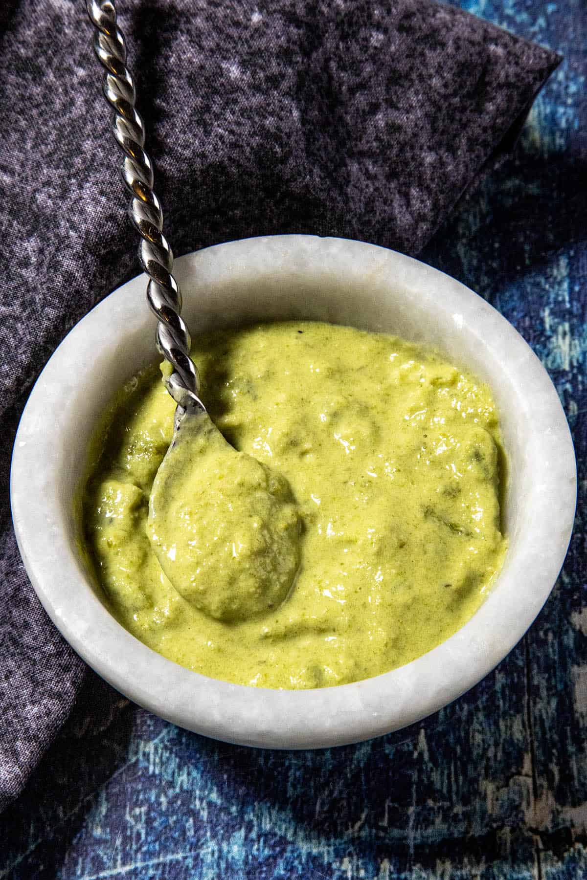 A marble bowl filled with thick green Spicy Roasted Jalapeño Sauce or chutney, with a twisted metal spoon resting inside, placed on a dark fabric napkin and a blue textured surface.