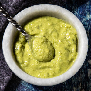 A marble bowl filled with thick, green Roasted Jalapeño Sauce. A metal spoon rests inside the bowl, which sits on a dark blue textured surface.
