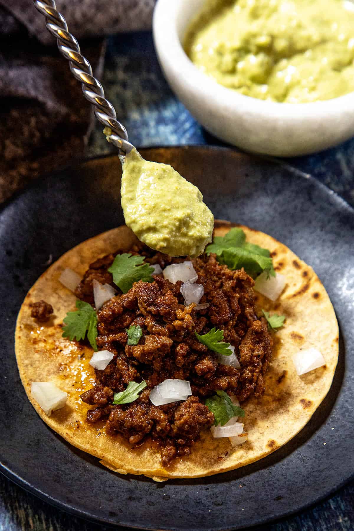 A street taco with spicy Roasted Jalapeño Sauce sits on a black plate, featuring a corn tortilla topped with seasoned ground meat, chopped onions, and cilantro. A spoonful of green guacamole is being held above it, ready to be added. In the background, there’s a bowl of guacamole waiting to complement the vibrant flavors.