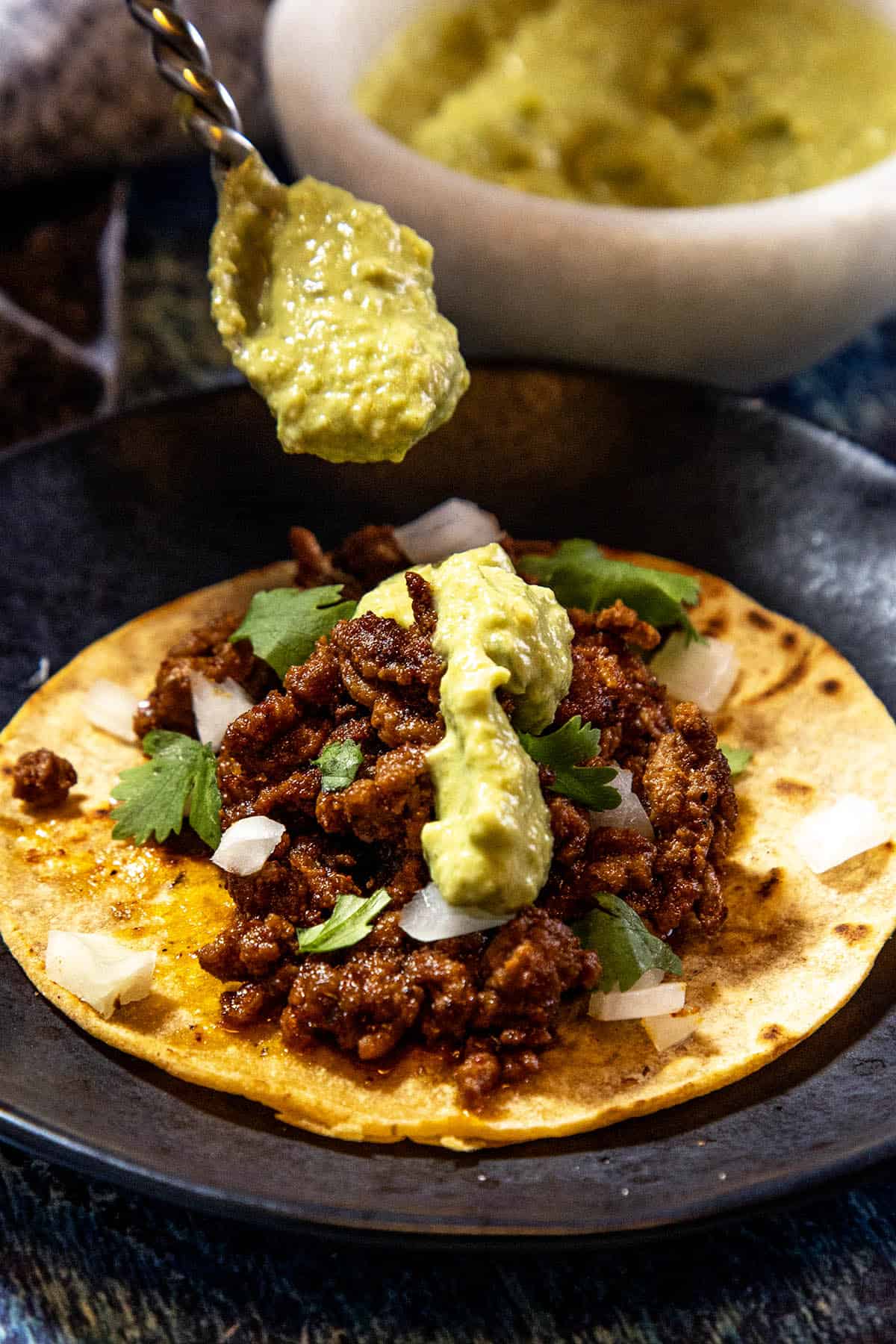 A crispy street taco with Roasted Jalapeño Sauce, topped with seasoned ground meat, chopped onions, cilantro, and a dollop of guacamole being spooned on, served on a dark plate.