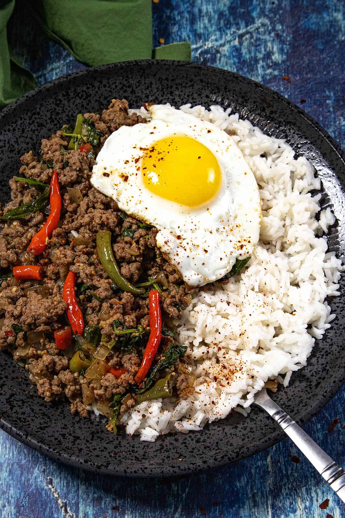 A black plate with white rice, Thai Basil Beef (Pad Kra Pao Neua) stir-fry made from ground beef, red chilies, and greens, topped with a fried egg, on a blue textured surface.