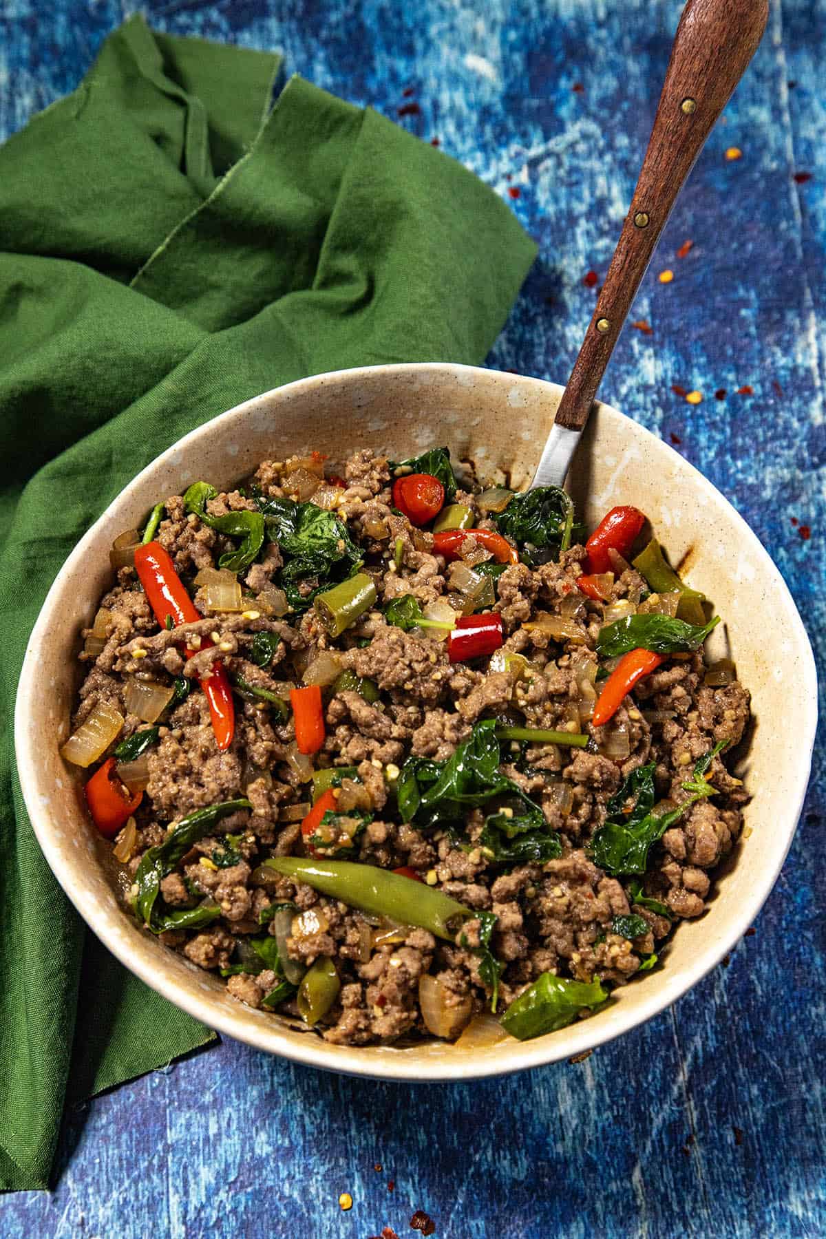 A serving bowl of Thai Basil Beef (Pad Kra Pao Neua) with colorful bell peppers, green vegetables, and spices, placed on a blue textured surface with a green napkin and a wooden-handled spoon.