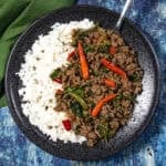 A black plate with white rice and spicy Thai Basil Beef (Pad Kra Pao Neua), topped with red chili peppers and vegetables, is placed on a blue surface with a fork and a green napkin nearby.