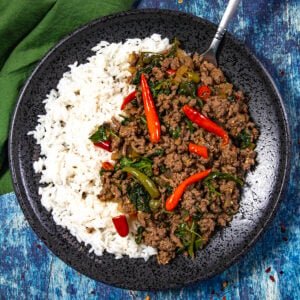 A black plate with white rice and spicy Thai Basil Beef (Pad Kra Pao Neua), topped with red chili peppers and vegetables, is placed on a blue surface with a fork and a green napkin nearby.