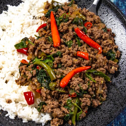 A black plate with white rice and spicy Thai Basil Beef (Pad Kra Pao Neua) stir-fry, topped with red chili peppers and greens, with a fork on the side.