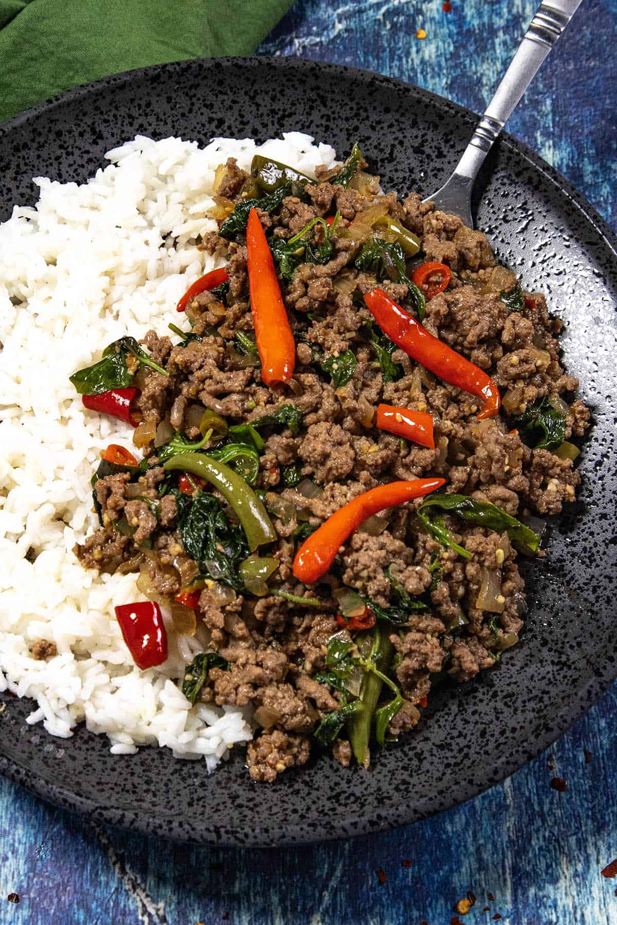 A black plate with white rice and spicy Thai Basil Beef (Pad Kra Pao Neua) stir-fry, topped with red chili peppers and greens, with a fork on the side.