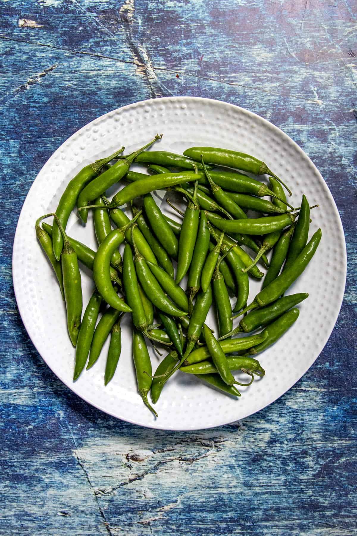 A white plate filled with fresh green Thai chilies sits on a textured blue surface.