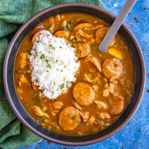 A bowl of rich Cajun Chicken and Sausage Gumbo with sliced sausage, chunks of meat, and vegetables is served with a mound of white rice on top.