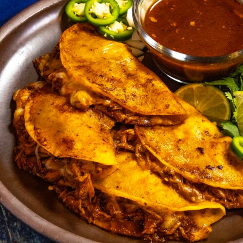 A plate loaded with four Chicken Birria Tacos (Birria de Pollo) served with a bowl of birria consome, with fresh lime, jalapeno slices, and cilantro for garnish
