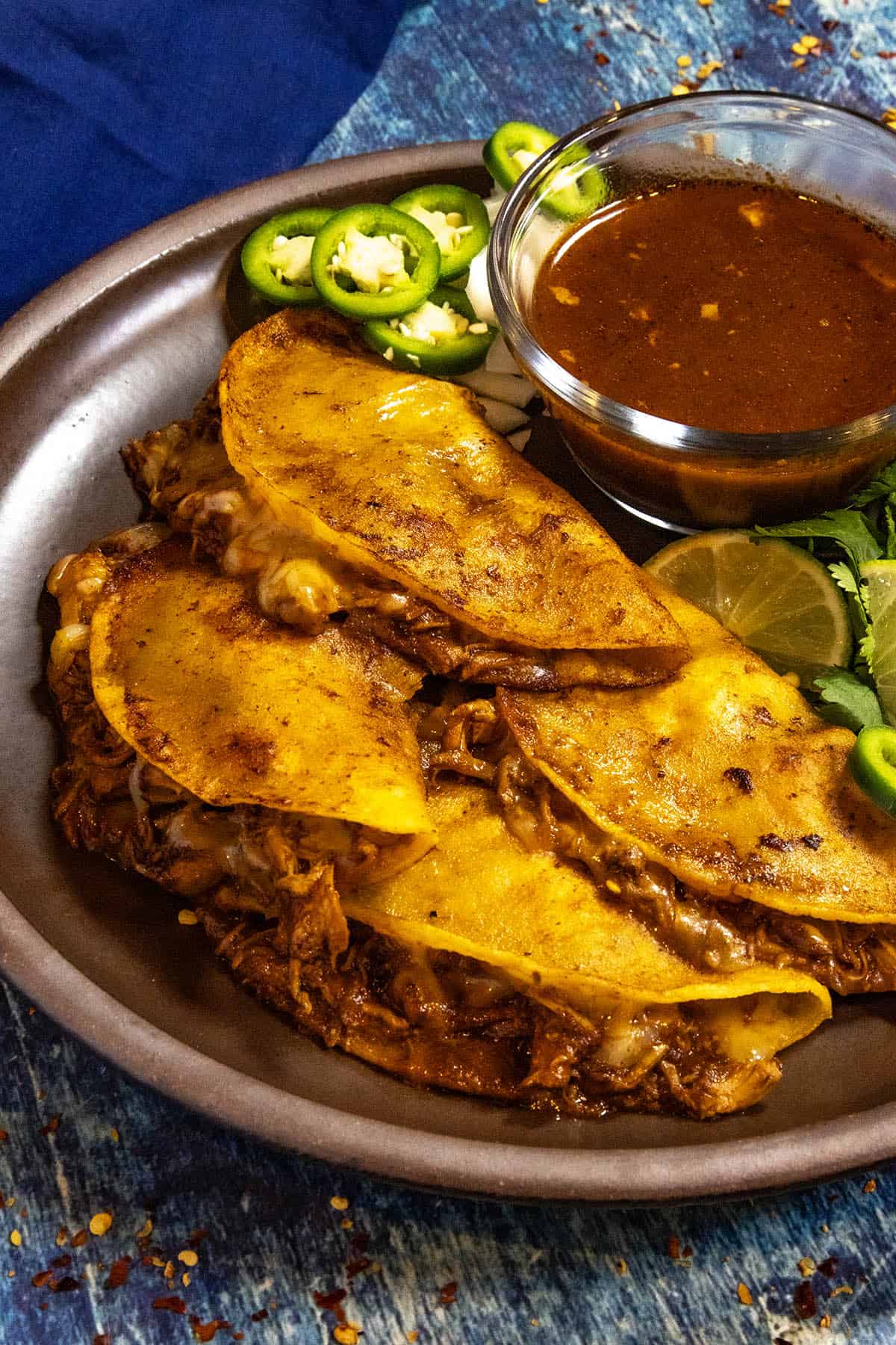 A plate loaded with four Chicken Birria Tacos (Birria de Pollo) served with a bowl of birria consome, with fresh lime, jalapeno slices, and cilantro for garnish