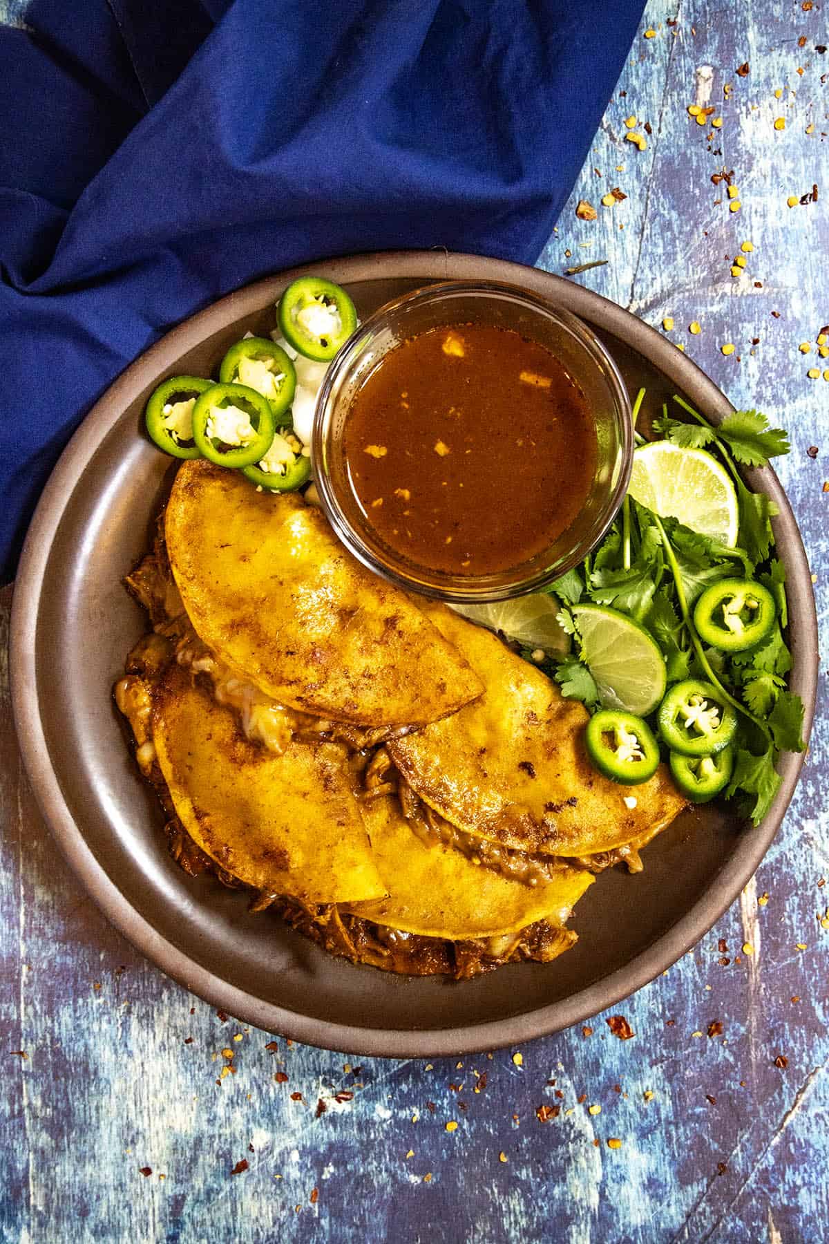 A plate stacked with four Chicken Birria Tacos (Birria de Pollo) served with fresh lime, jalapeno slices, cilantro, and consome